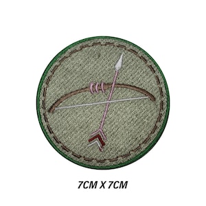 May include: Circular embroidered patch with a bow and arrow design. The patch has a green border and a light green background. The bow is brown, and the arrow is pink with a red fletching. The patch measures 7cm x 7cm.