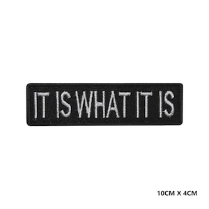 May include: Black embroidered patch with white text that reads "IT IS WHAT IT IS". The patch is 10 cm by 4 cm.