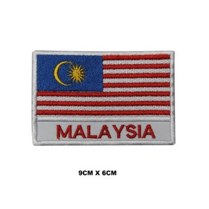 Op de afbeelding: Een geborduurde patch met de vlag van Maleisië met een gele halve maan en ster op een blauwe achtergrond, 14 rode en witte strepen en het woord "MALAYSIA" in witte letters op een grijze achtergrond. De patch meet 9 cm bij 6 cm.