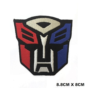 May include: A black, red, white and blue embroidered iron-on patch featuring the Transformers logo. The logo is a stylised robot face with a black background and red and blue accents. The patch measures 8.8 cm by 8 cm.