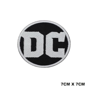 May include: A circular embroidered patch featuring the white letters DC centered on a black background with a white border. The patch measures 7 cm by 7 cm. The design consists of bold, block-style typography within a circular frame, showcasing a high-contrast black and white color scheme throughout the entire embroidered textile piece.