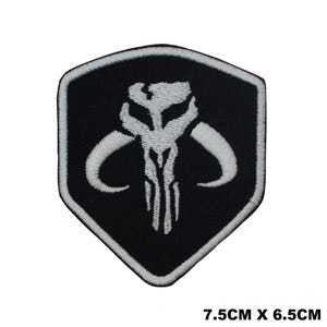 May include: A shield-shaped embroidered patch featuring a white Mythosaur skull design on a black background with a white border. The patch measures 7.5 cm by 6.5 cm, as indicated by the text printed in the bottom right corner of the image.