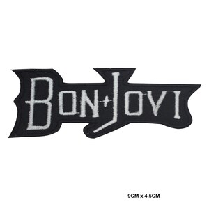 May include: A black embroidered patch featuring the white text BON JOVI in a stylised font. The patch has a jagged, angular shape and measures 9 cm by 4.5 cm. The embroidery is dense, creating a clear contrast between the black background and white lettering.