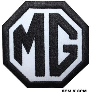 Könnte beinhalten: Schwarz-weißer, gestickter Aufnäher mit den Buchstaben "MG" in einem achteckigen Rahmen. Misst 8 cm x 8 cm. Das Design zeigt eine kühne, stilisierte Schrift.