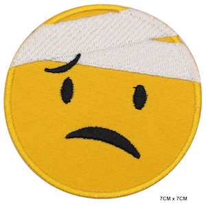Op de afbeelding: Gele geborduurde strijkpatch met een verdrietig gezicht met een wit verband op zijn voorhoofd. De patch is 7 cm bij 7 cm.