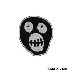 May include: An embroidered patch featuring a black, rounded, mask-like face with white stitched eyes and a white stitched mouth showing teeth. The patch has a white border and measures 6cm x 7cm as indicated by the text in the image.