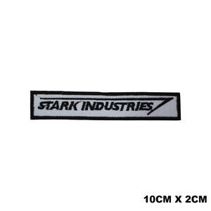 以下が含まれることがあります： 白い背景に黒い縁取り、黒い文字でSTARK INDUSTRIESと刺繍された長方形のワッペン。右側には黒い矢印のようなグラフィックがデザインされています。ワッペンのサイズは10cm x 2cmです。