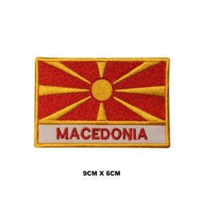 May include: Embroidered iron-on patch featuring the flag of North Macedonia. The flag is red with a yellow sun with eight rays. The patch is 9 cm by 6 cm and has the word "MACEDONIA" written in yellow below the flag.