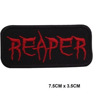 Puede incluir: Un parche rectangular bordado con fondo negro y la palabra REAPER cosida en hilo rojo. El parche presenta un borde negro grueso y mide 7,5 cm por 3,5 cm, tal como indica el texto impreso debajo del objeto.