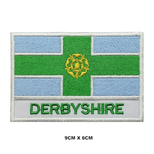 May include: An embroidered patch featuring the flag of Derbyshire, a white background with a green cross and a gold rose in the centre. The text "DERBYSHIRE" is written in white below the flag. The patch measures 9 cm by 6 cm.