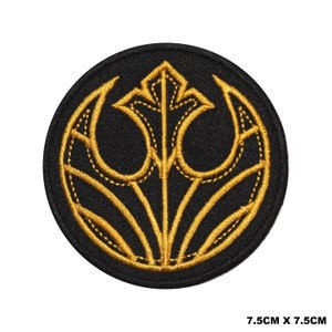 May include: A circular embroidered patch featuring a black background with a gold-coloured stylised emblem design. The emblem consists of a central vertical line with symmetrical curved branches. The patch measures 7.5 cm by 7.5 cm in size.