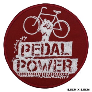 Könnte beinhalten: Runder roter gestickter Aufnäher mit einem weißen Fahrrad und einer erhobenen Faust. Der Text "PEDAL POWER" ist in weiß unter dem Fahrrad geschrieben.