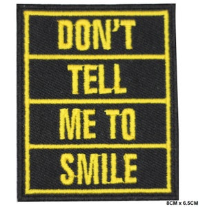 May include: Black iron-on patch with yellow embroidered text that reads "Don't Tell Me To Smile". The patch measures 8 cm by 6.5 cm.