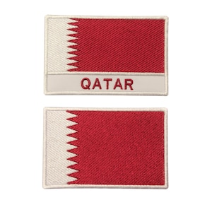 Puede incluir: Dos parches bordados blancos con un diseño de bandera roja y blanca. El parche superior tiene la palabra "QATAR" escrita en blanco sobre un fondo gris. El parche inferior muestra la bandera de Qatar.