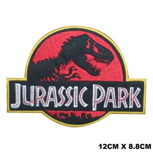 May include: An embroidered patch featuring the Jurassic Park logo with a black T-Rex silhouette against a red circular background, bordered in gold. The text JURASSIC PARK is in white with red outlines on a black rectangular base, measuring 12cm x 8.8cm.
