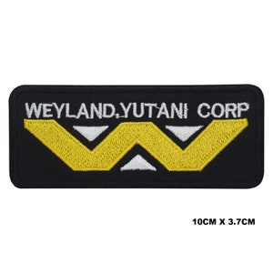 Peut inclure: Écusson rectangulaire noir avec le texte "WEYLAND, YUTANI CORP" en blanc. L'écusson présente un logo jaune et blanc. Il mesure 10 cm sur 3,7 cm.