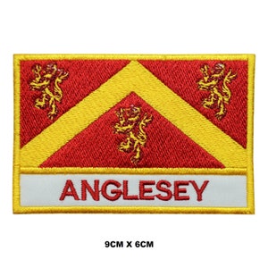 May include: A red and yellow embroidered patch with three gold lions and the word "ANGLESEY" in yellow lettering on a white background. The patch measures 9 cm by 6 cm.