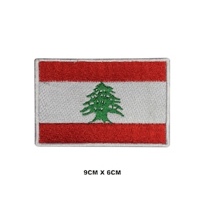 Może przedstawiać: Haftowana naszywka z flagą Libanu. Prostokątna naszywka ma czerwone paski u góry i u dołu, z białym paskiem pośrodku. Zielony cedr jest wyśrodkowany na białym pasku. Naszywka ma wymiary 9 cm x 6 cm.