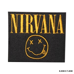 Puede incluir: Un parche rectangular negro bordado con la palabra NIRVANA en hilo dorado sobre una cara sonriente dorada bordada con ojos en forma de X y la lengua fuera. El parche mide 8,3 cm por 7,4 cm como se indica en la imagen.