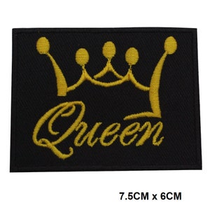Może przedstawiać: Czarna naszywka do prasowania z haftowaną złotą koroną i napisem "Queen". Naszywka ma wymiary 7,5 cm x 6 cm.