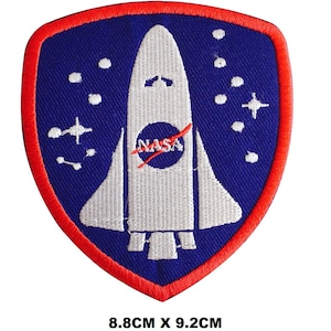 Puede incluir: Un parche bordado azul y rojo con una lanzadera espacial blanca y el logotipo de la NASA. El parche mide 8,8 cm por 9,2 cm.