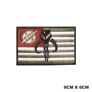 May include: A rectangular embroidered patch measuring 9cm x 6cm featuring a flag design with horizontal olive green and white stripes. A red canton contains a circular emblem with a white skull symbol. A black Mandalorian helmet icon is centered.