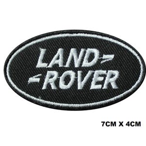 May include: A black oval patch featuring the white embroidered text "LAND ROVER". The patch is edged with white stitching. The dimensions are 7cm x 4cm.