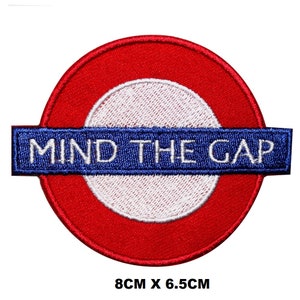 May include: Red and white embroidered patch with the text "MIND THE GAP" in blue. The patch is 8 cm by 6.5 cm.