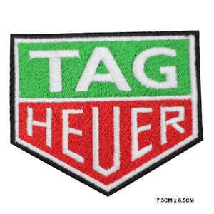Può includere: Toppa ricamata a forma di scudo con la scritta "TAG HEUER" in bianco su sfondo verde e rosso. La toppa ha un bordo nero e misura 7,5 cm x 6,5 cm.