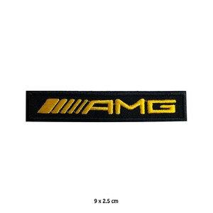 Puede incluir: Un parche rectangular negro con el texto amarillo "AMG". El parche mide 9 x 2,5 cm. El logotipo de AMG es un diseño estilizado, comúnmente asociado con vehículos de alto rendimiento.