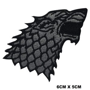Puede incluir: Un parche bordado con forma de perfil de cabeza de lobo, que presenta hilo negro y gris con un patrón texturizado similar a escamas. El diseño incluye un borde dentado y la boca abierta. Las dimensiones indicadas son 6 cm x 5 cm.