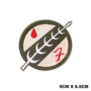 Op de afbeelding: Een ronde geborduurde patch van 9 cm bij 6,5 cm. Het ontwerp heeft een crèmekleurige achtergrond met een donker olijfgroene diagonale streep, een wit speerachtig icoon, een rode druppelvorm en een rood gestileerd teken dat lijkt op een 7.