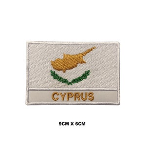 May include: White embroidered patch with the outline of Cyprus in gold and a green laurel wreath. The text "CYPRUS" is written in gold below the outline. The patch measures 9 cm by 6 cm.
