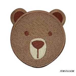 以下が含まれることがあります： 茶色のクマの顔の刺繍ワッペン、7cm x 6.5cm。クマは茶色の耳、目、鼻を持ち、クリーム色の鼻をしています。このワッペンは、衣類やアクセサリーへの貼り付け用にデザインされています。
