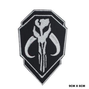 May include: A black and white embroidered patch featuring a stylised skull design with curved horns. The patch is shaped like a shield with a pointed bottom and measures 9cm x 6cm. The white thread creates a detailed graphic on a black background.