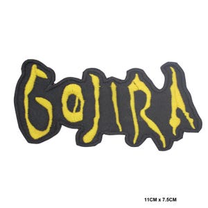 Op de afbeelding: Een geborduurde patch met het woord GOJIRA in geel garen op een zwarte achtergrond. De letters hebben een grillig, gestileerd lettertype. De patch meet 11 cm bij 7,5 cm, zoals aangegeven door de tekst in de rechterbenedenhoek.