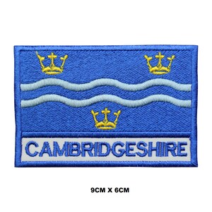 Pode incluir: Etiqueta bordada com a bandeira de Cambridgeshire, um fundo azul com três coroas douradas e duas linhas onduladas brancas. O texto "CAMBRIDGESHIRE" está escrito em branco abaixo da bandeira. A etiqueta mede 9 cm por 6 cm.