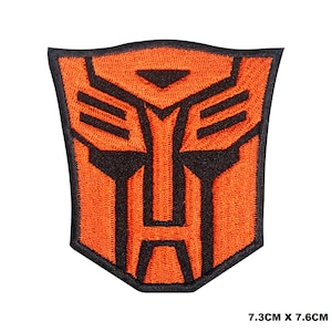 May include: Orange and black embroidered iron-on patch featuring the Transformers logo. The logo is a stylised robot face with a triangular head and a rectangular body. The patch measures 7.3 cm by 7.6 cm.
