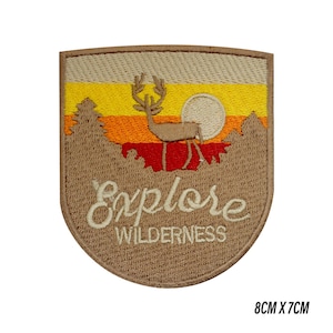 Op de afbeelding: Geborduurde patch met een schildvorm, met een herten silhouet, bomen en een zonsondergang ontwerp. De tekst "Explore Wilderness" is in cursief geborduurd. De patch meet 8 cm bij 7 cm.
