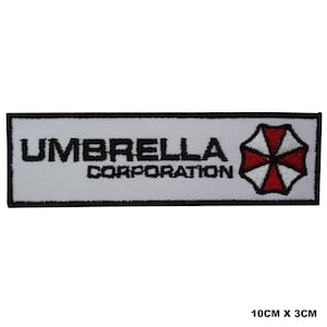 May include: A rectangular embroidered patch with a white background and a black border. It features the black text UMBRELLA CORPORATION and a red and white umbrella-shaped logo on the right side. The patch measures 10 cm by 3 cm.