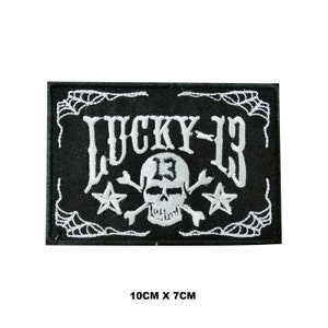 Könnte beinhalten: Schwarz-weiß gestickter Patch mit Totenkopf und Knochen, zwei Sternen und dem Text "LUCKY-13". Der Patch ist 10 cm x 7 cm groß.