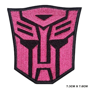 May include: Pink and black embroidered iron-on patch featuring the Transformers logo. The logo is a stylized robot face with a black outline and a pink interior. The patch measures 7.3 cm by 7.6 cm.