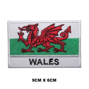 May include: A white embroidered patch featuring a red dragon and the text "WALES" in black. The patch measures 9 cm by 6 cm.