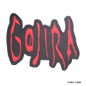 May include: A black embroidered patch featuring the word GOJIRA in a stylized, jagged red font. The patch has a black border and measures 11 cm by 7.5 cm, as indicated by the text printed in the bottom right corner of the image.