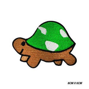 Könnte beinhalten: Gestickter Schildkrötenaufnäher mit grünem Panzer und weißen Punkten sowie einem braunen Körper. Der Aufnäher misst 8 cm x 6 cm.