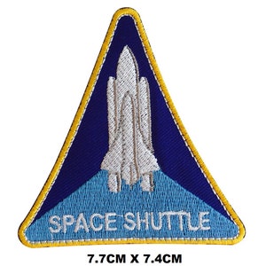May include: A blue and white embroidered patch featuring a space shuttle design. The patch is triangular with a yellow border and the text "SPACE SHUTTLE" in white. The patch measures 7.7 cm by 7.4 cm.