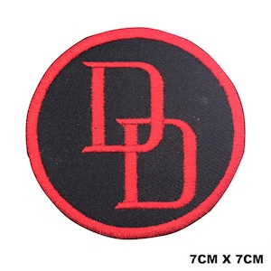 May include: A circular embroidered patch featuring a black background with a red border and a red double D logo in the center. The patch measures 7 cm by 7 cm as indicated by the text printed in the lower right corner of the image.