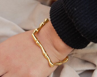May include: A gold-colored square bangle bracelet with a bamboo-like design. The bracelet is worn on a wrist, and the image is a close-up shot. The bracelet is made of metal and has a shiny finish.