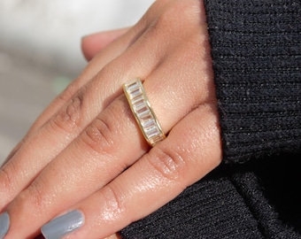 May include: A gold-colored ring with rectangular, clear stones set in a row. The ring is on a finger, with a black sweater in the background. The ring is a band style.