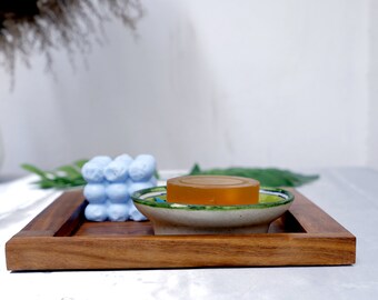 May include: A wooden tray with a light blue soap, a small ceramic dish with a yellow soap bar, and green leaves.